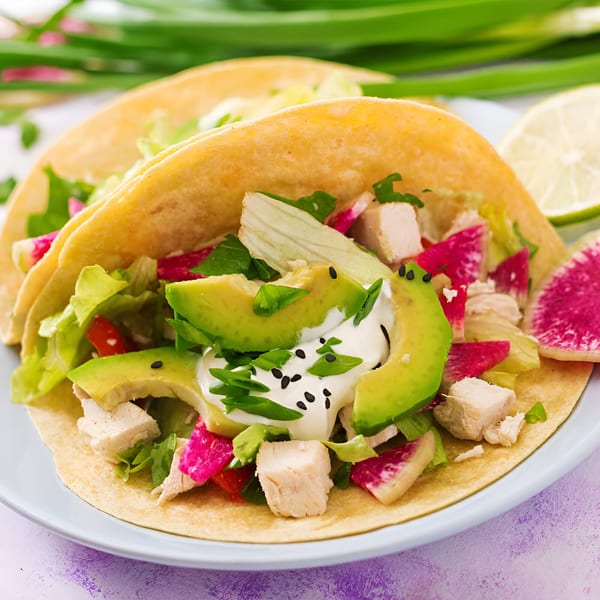 Two Los Amigos Mexican chicken tacos with avocado, radish, lettuce, and sour cream on a plate.